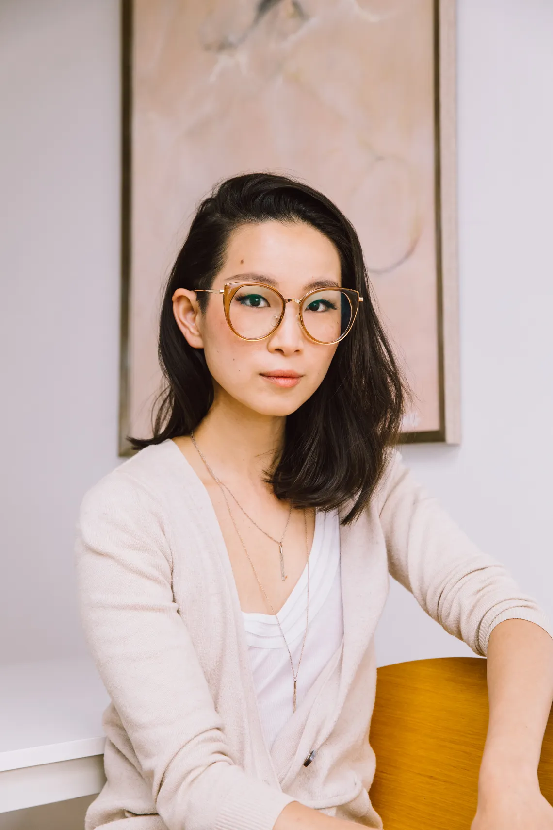 Tria Wen, wearing beige glasses and an off-white cardigan, seated before a light peach painting